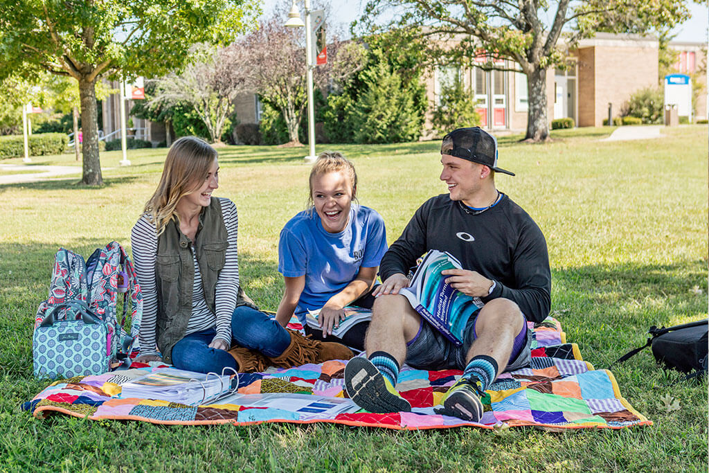 three students sitting on a blanket outside laughing