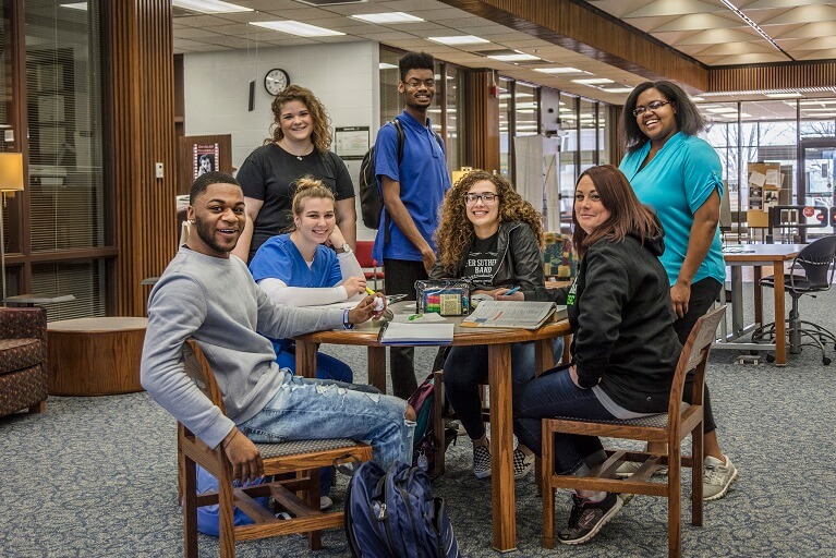 students sitting at a table in a library smiling at the camera