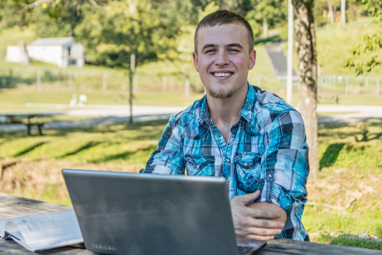 male student sitting on a picnic bench outside with a silver laptop