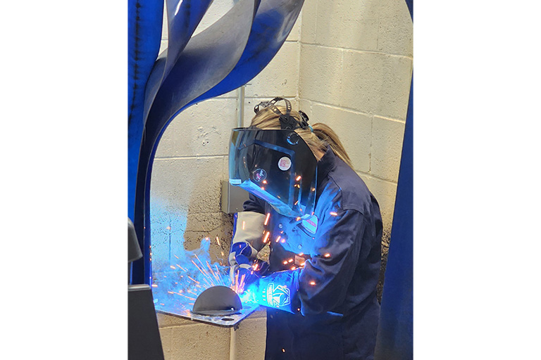 SCC student welding with a mask on while sparks and blue light shines on the welding coat that the student is wearing.